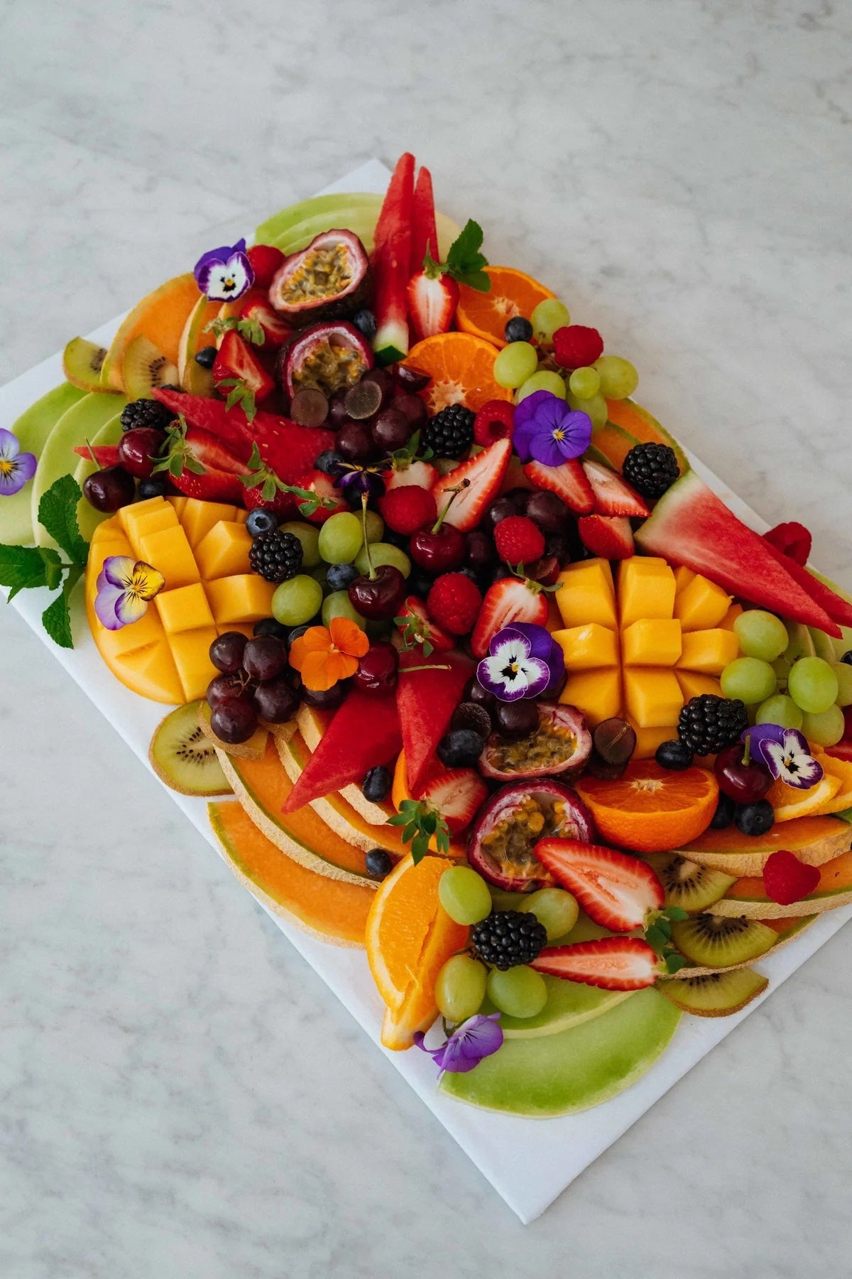 Vibrant cut fruit platter featuring mango, strawberries, oranges, and decorative edible flowers, perfect for sharing.