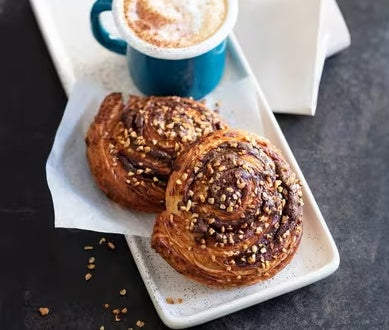 Chocolate brownie pastry with hazelnuts paired with a creamy coffee, served on a white plate.
